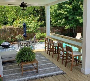 Covered backyard patio with seating, striped rug, hanging fan, and lush green trees beyond the fence