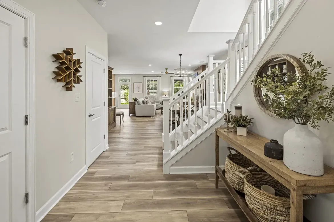 Bright hallway with wood flooring, white staircase, and console table with wreath décor leading to a living area