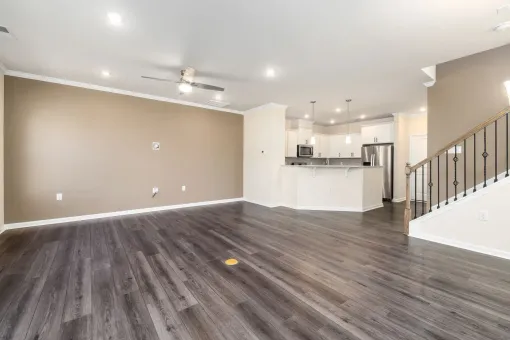 Empty modern living room with gray wood floors, beige walls, white kitchen, and staircase.