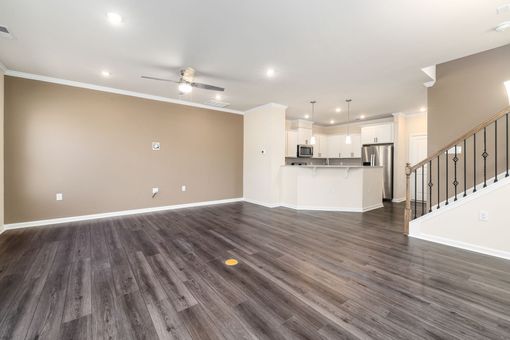 Empty modern living room with gray wood floors, beige walls, white kitchen, and staircase.