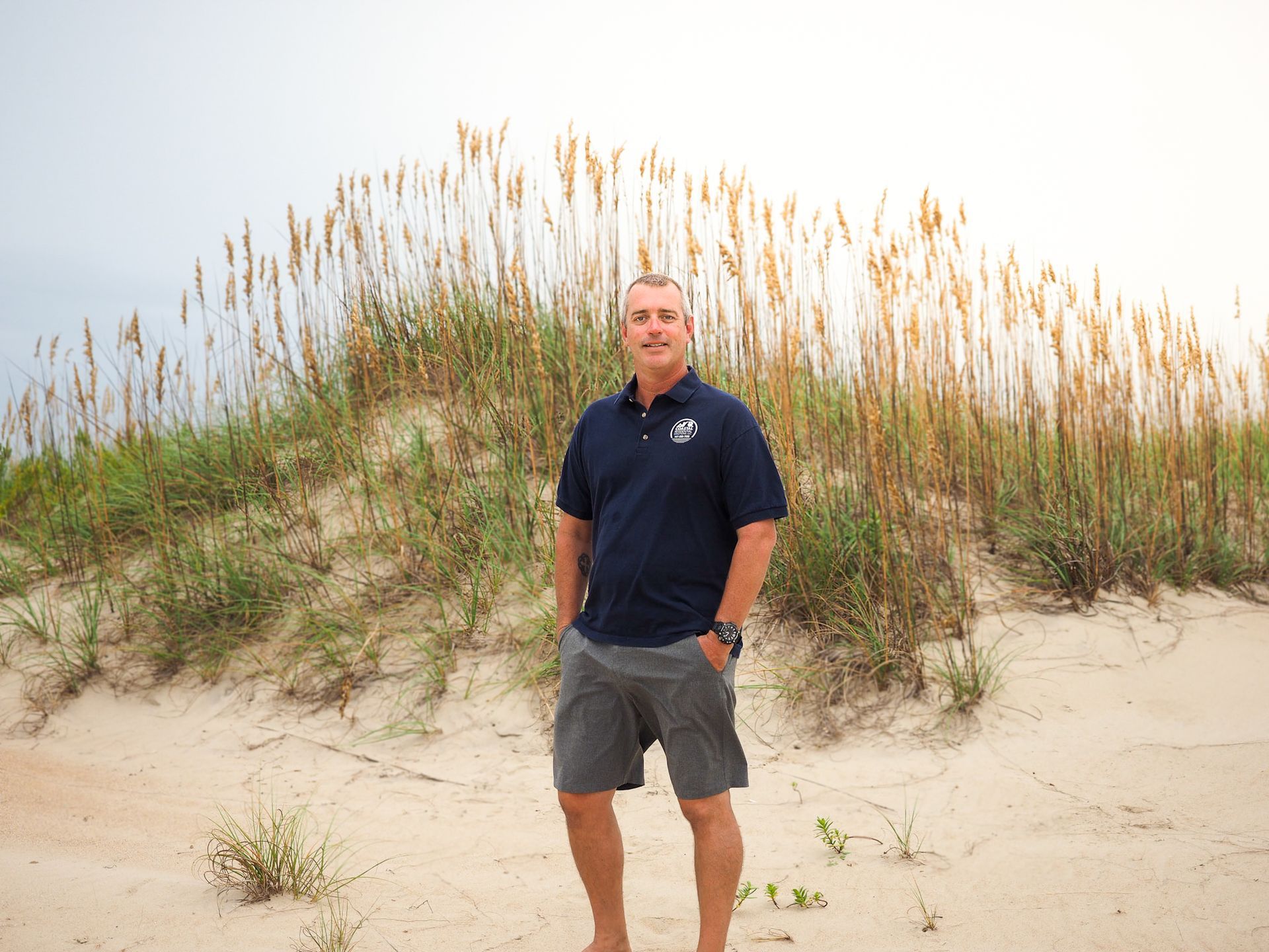 Man standing on sandy dunes in front of tall beach grass, wearing a dark polo and gray shorts.