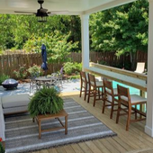 Covered backyard patio with sofa, striped rug, bar counter stools, and lush garden beyond