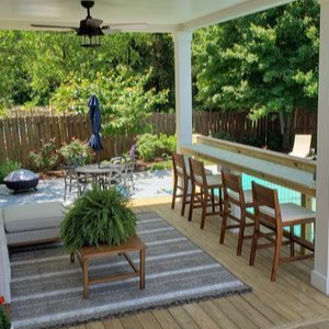 Covered backyard patio with sofa, striped rug, bar counter stools, and lush garden beyond