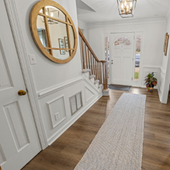 Bright hallway with white walls, wood floor, round mirror, staircase, and a runner rug leading to a front door.