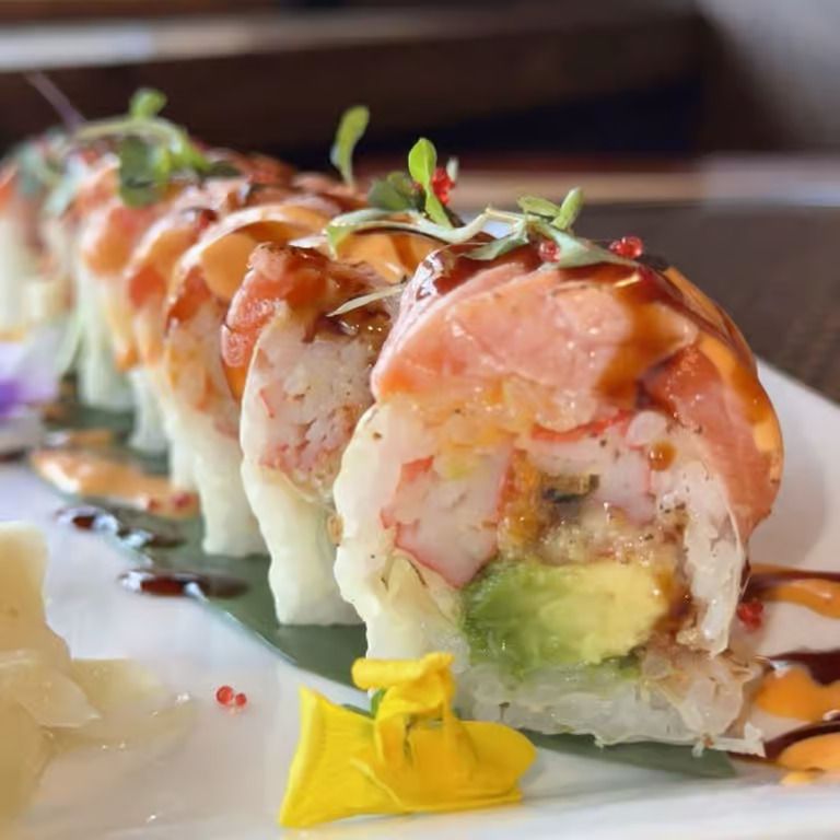 Sushi roll topped with salmon, avocado, and drizzled sauce on a white plate