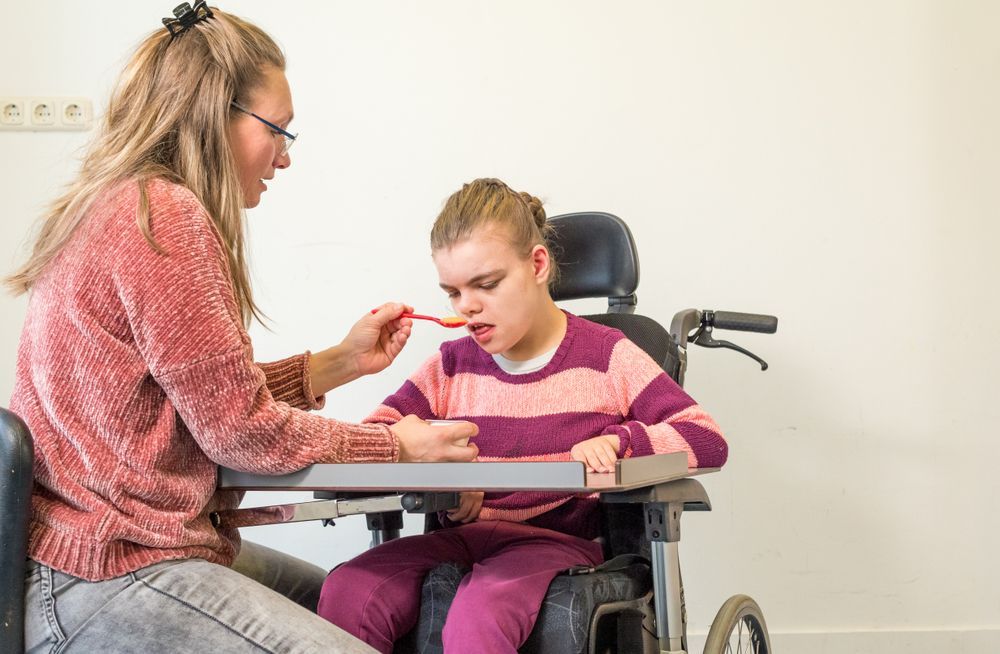 A Woman is Feeding a Young Girl in a Wheelchair With a Spoon — Collis Care In Dundowran, QLD