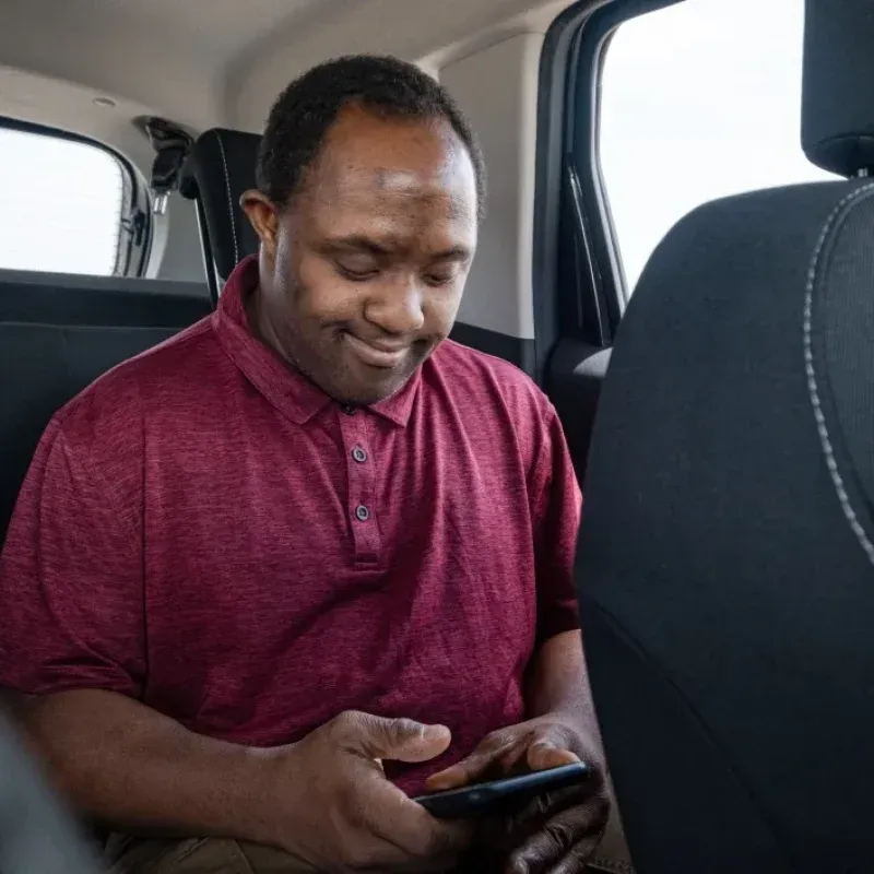 A Man in a Red Shirt is Sitting in the Back Seat of a Car Looking at His Phone — Collis Care In Dundowran, QLD