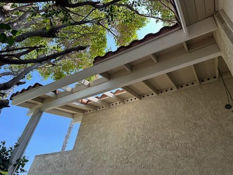 Beige covered patio with terracotta tile roof and textured stucco wall; tree branches overhead.