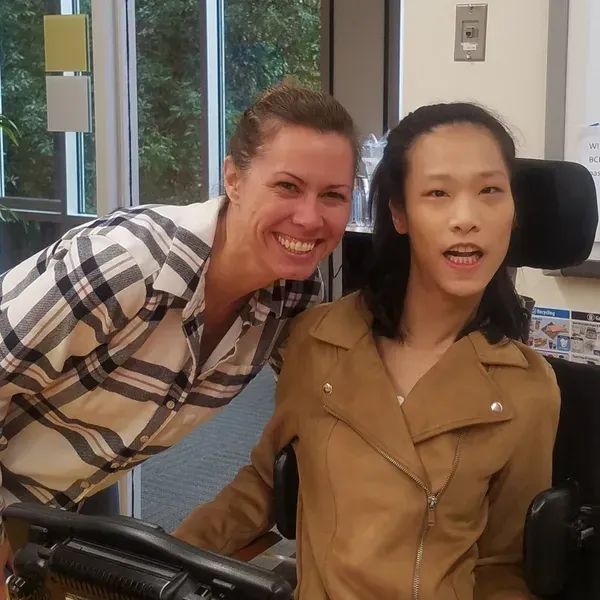 Woman smiles next to a person in a wheelchair. Both look at the camera, inside a room.