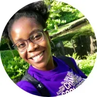 Person smiling outdoors, wearing glasses and a purple shirt, with trees and water in the background.