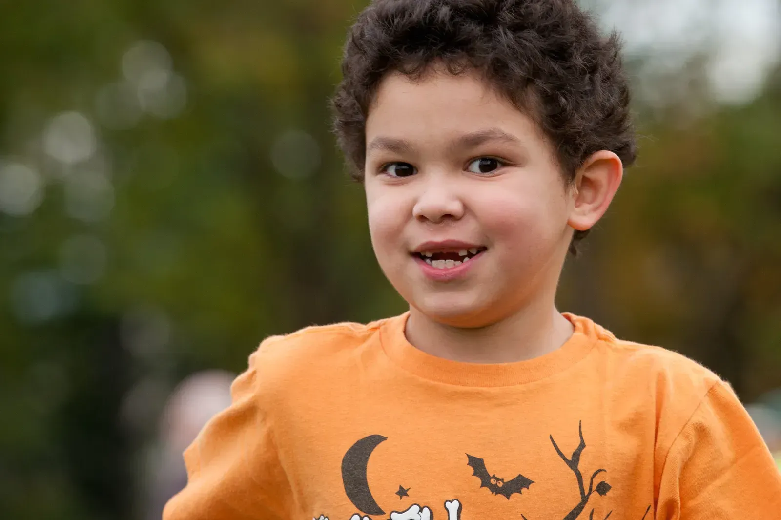 Boy in orange shirt with Halloween design smiles outdoors.