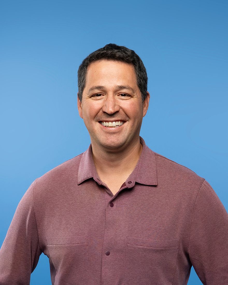Man smiling, wearing a plum-colored button-up shirt, with blue background. 