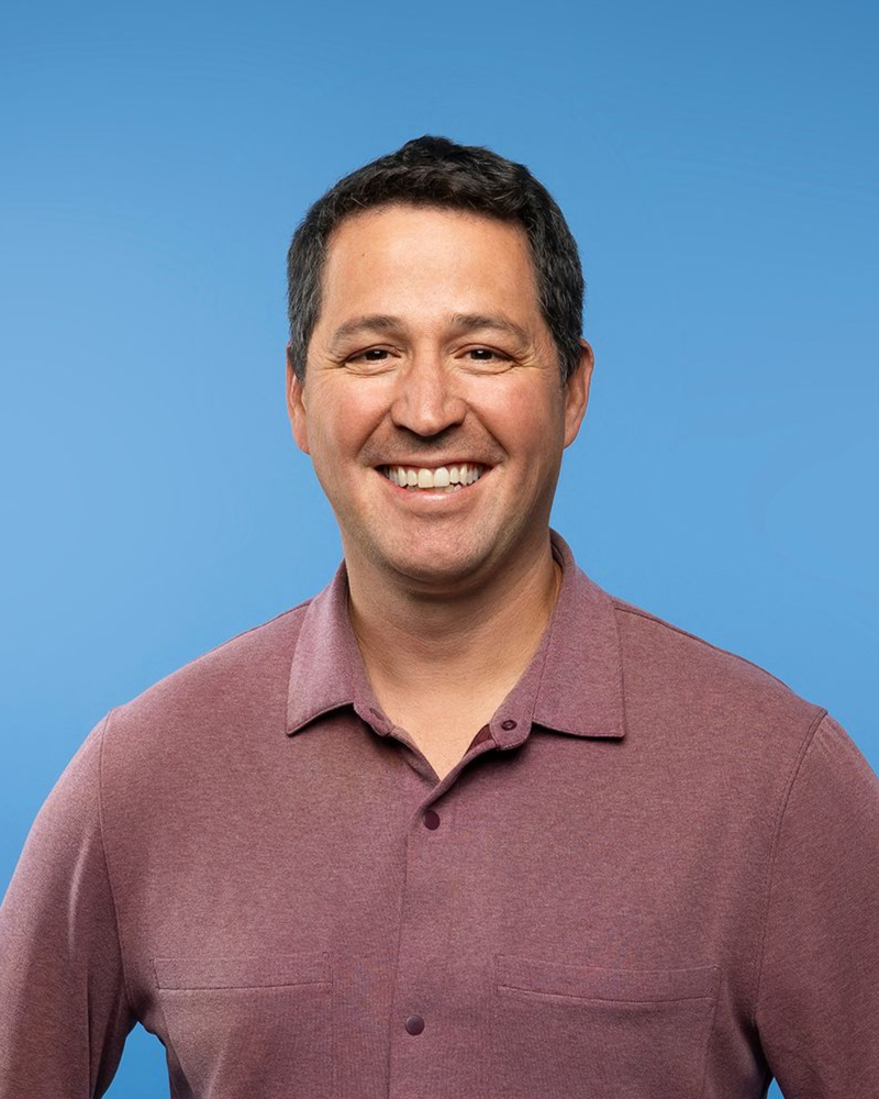 Man smiling, wearing a plum-colored button-up shirt, with blue background. 