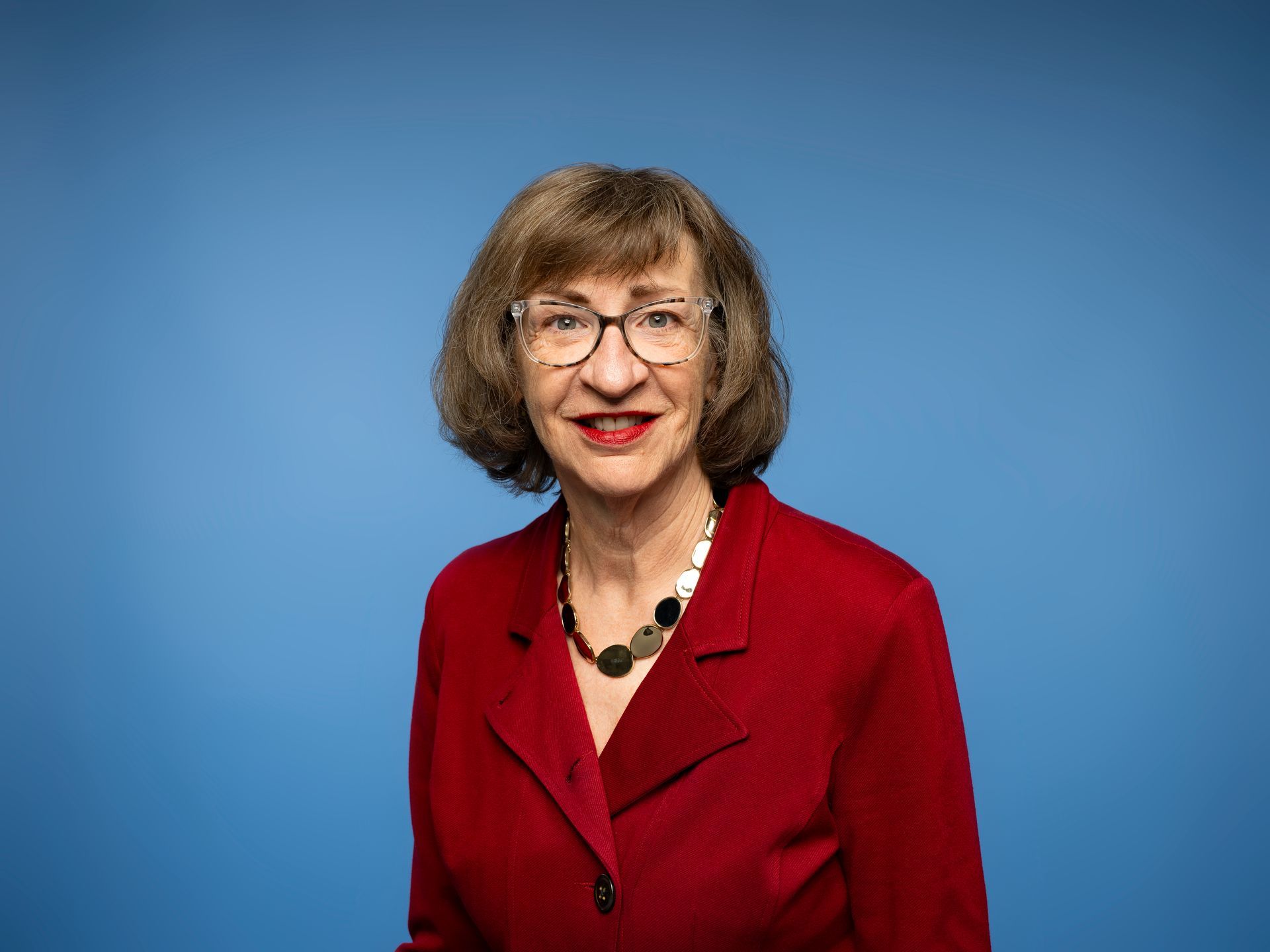 Woman with glasses and short brown hair smiles, wearing a beaded necklace; wood art in background.