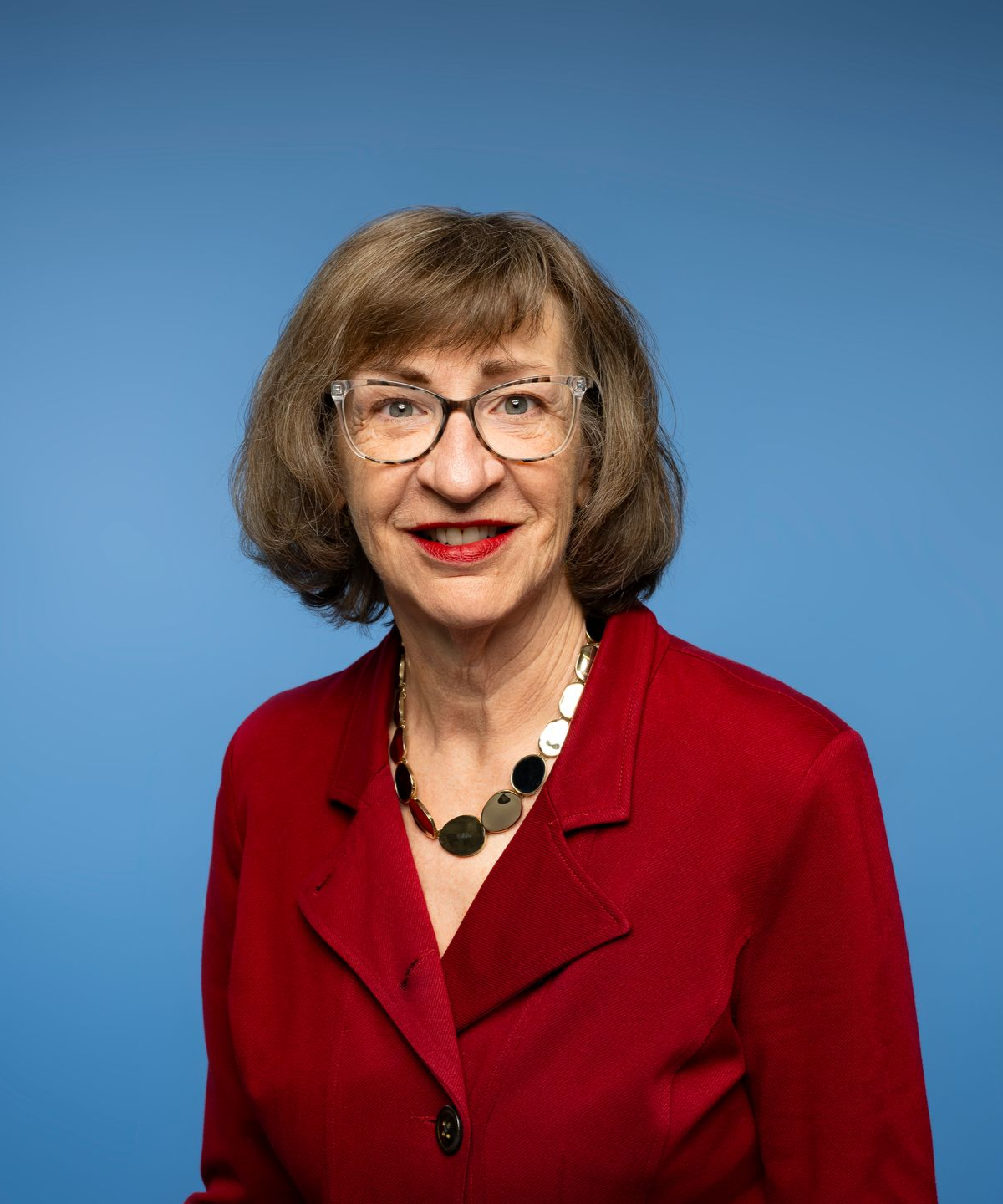 Woman with glasses and short brown hair smiles, wearing a beaded necklace; wood art in background.