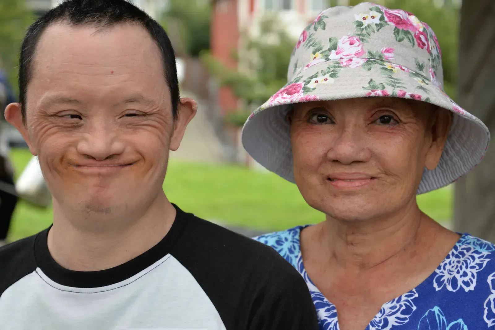 Man with Down syndrome smiles next to woman wearing floral hat, outdoors.