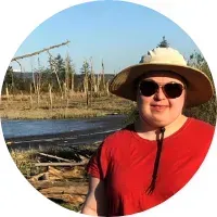 Person in red shirt and hat outdoors with sunglasses, trees and water in the background.