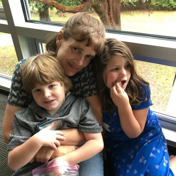 Woman with two children, posing in front of a window. One child is eating, the other is holding a bag.