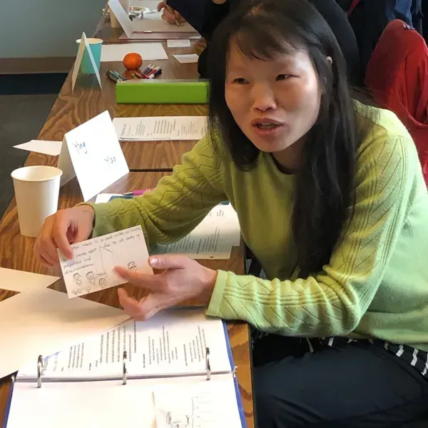 Woman in green sweater holds a card while seated at a table with papers, cups, and a binder.
