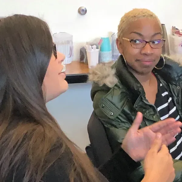 Two women in conversation. One with blonde hair and glasses gesturing. The other with long brown hair, talking. Indoors.