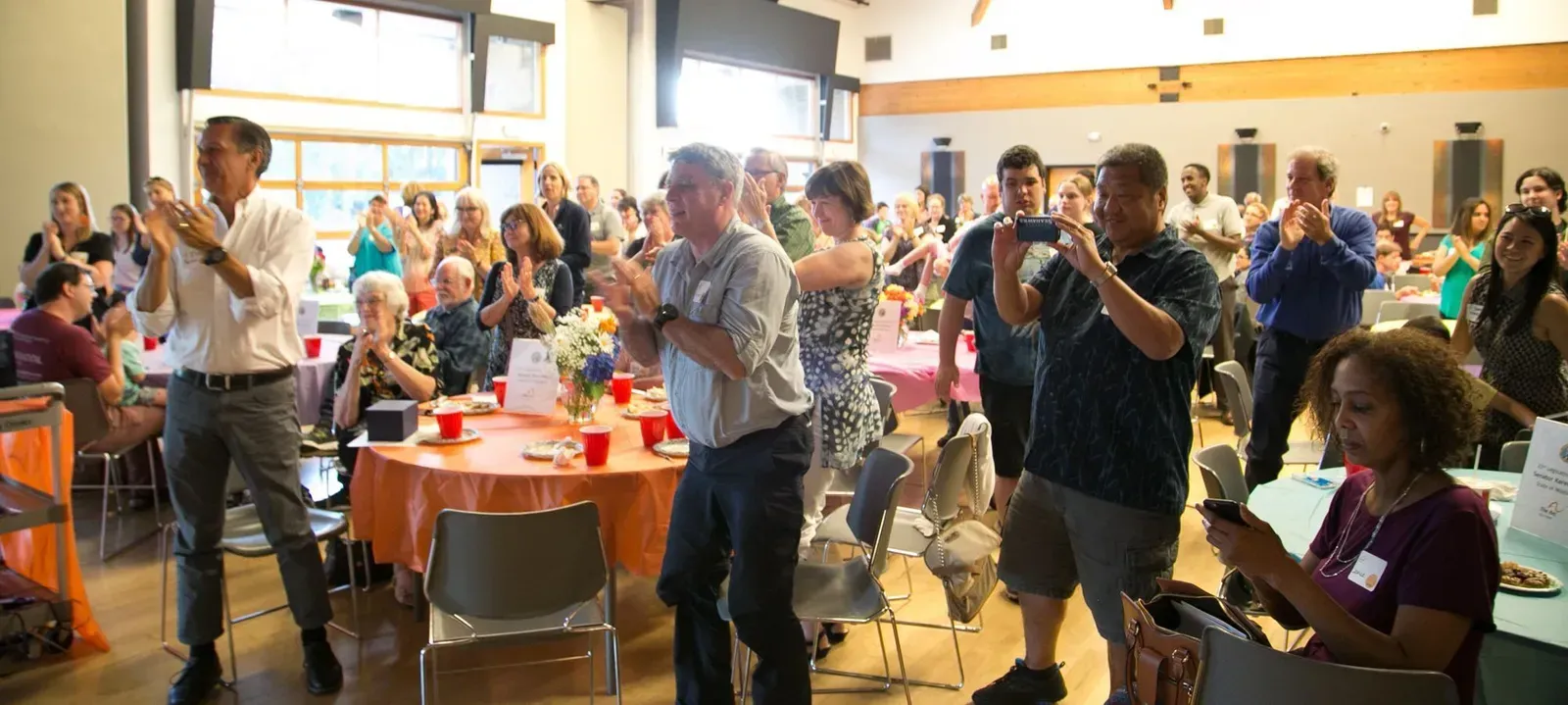 People clapping in a large hall with tables set for an event.