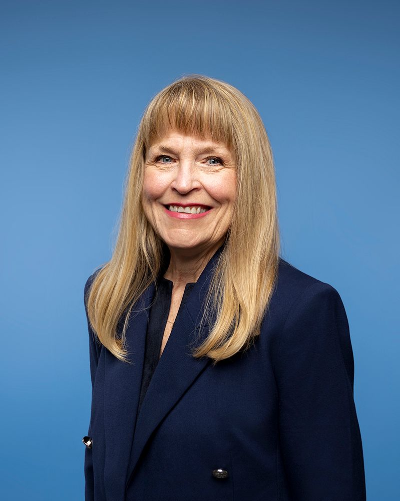 Woman with blonde hair, wearing a yellow top, smiling slightly, in front of a neutral-colored background.
