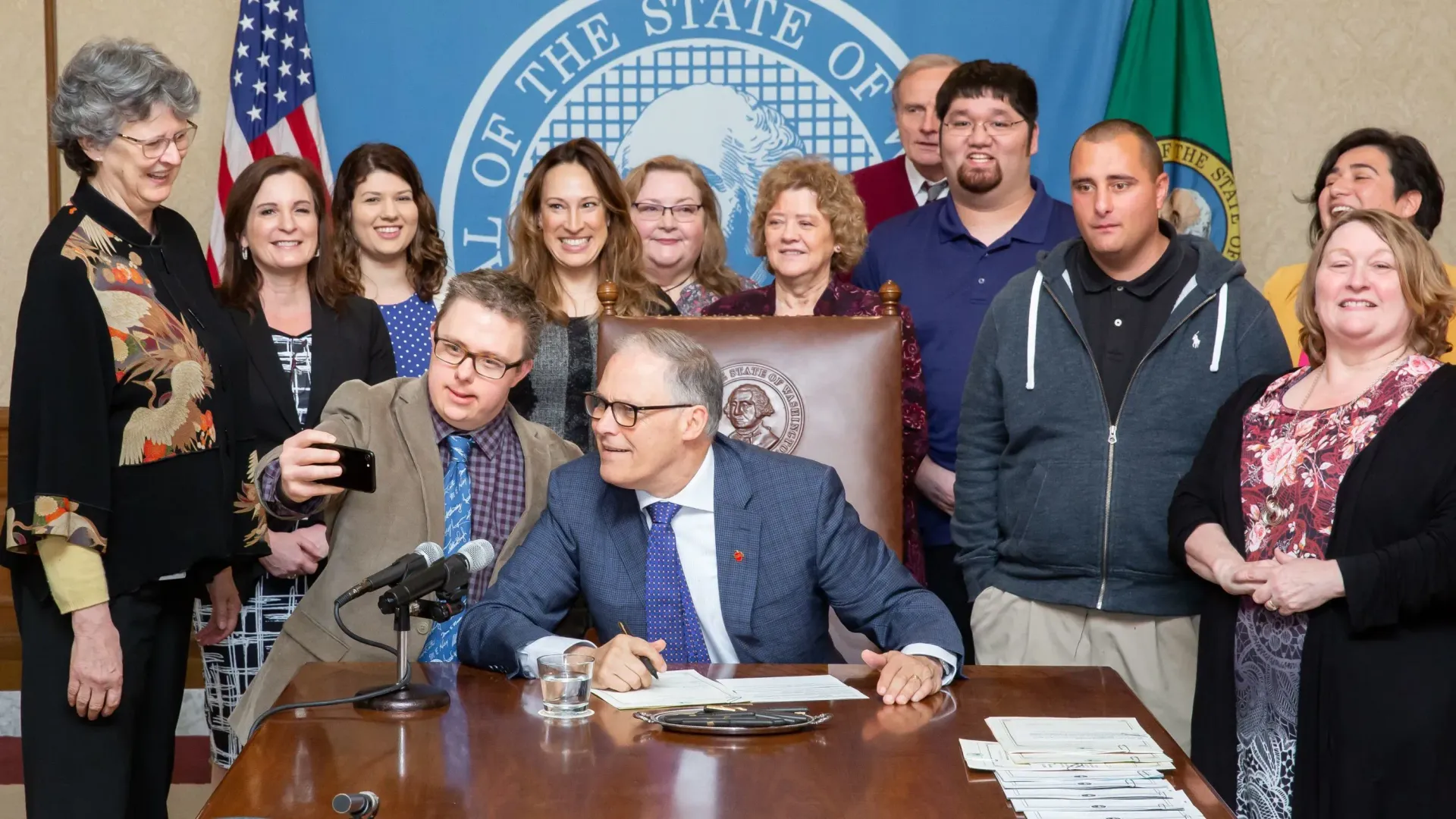 People gather around the governor signing documents at a desk; another takes a selfie. State seal in the background.