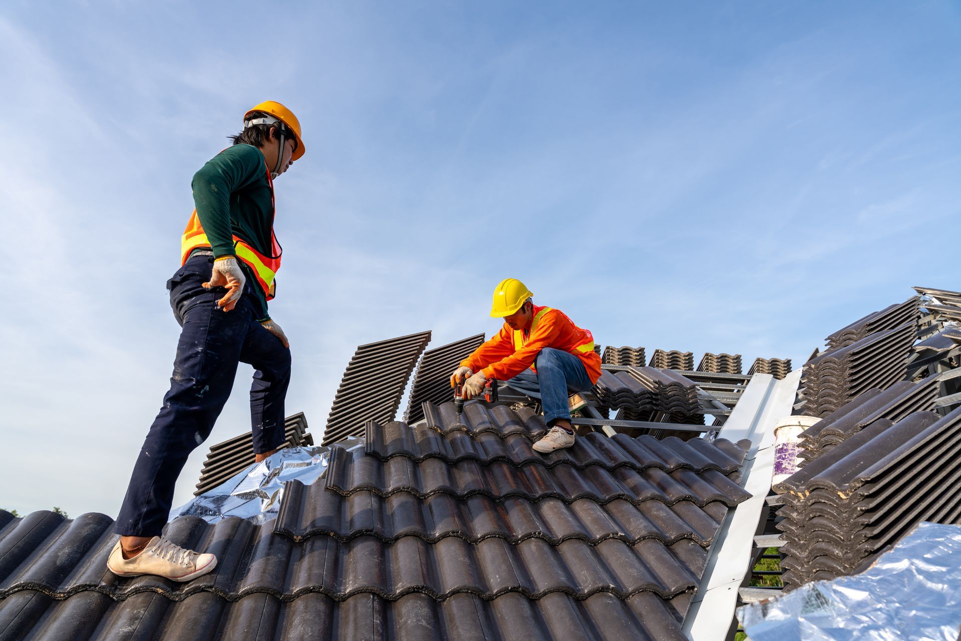 Two roofers on a rooftop installing dark tiles, wearing safety gear, under a blue sky.