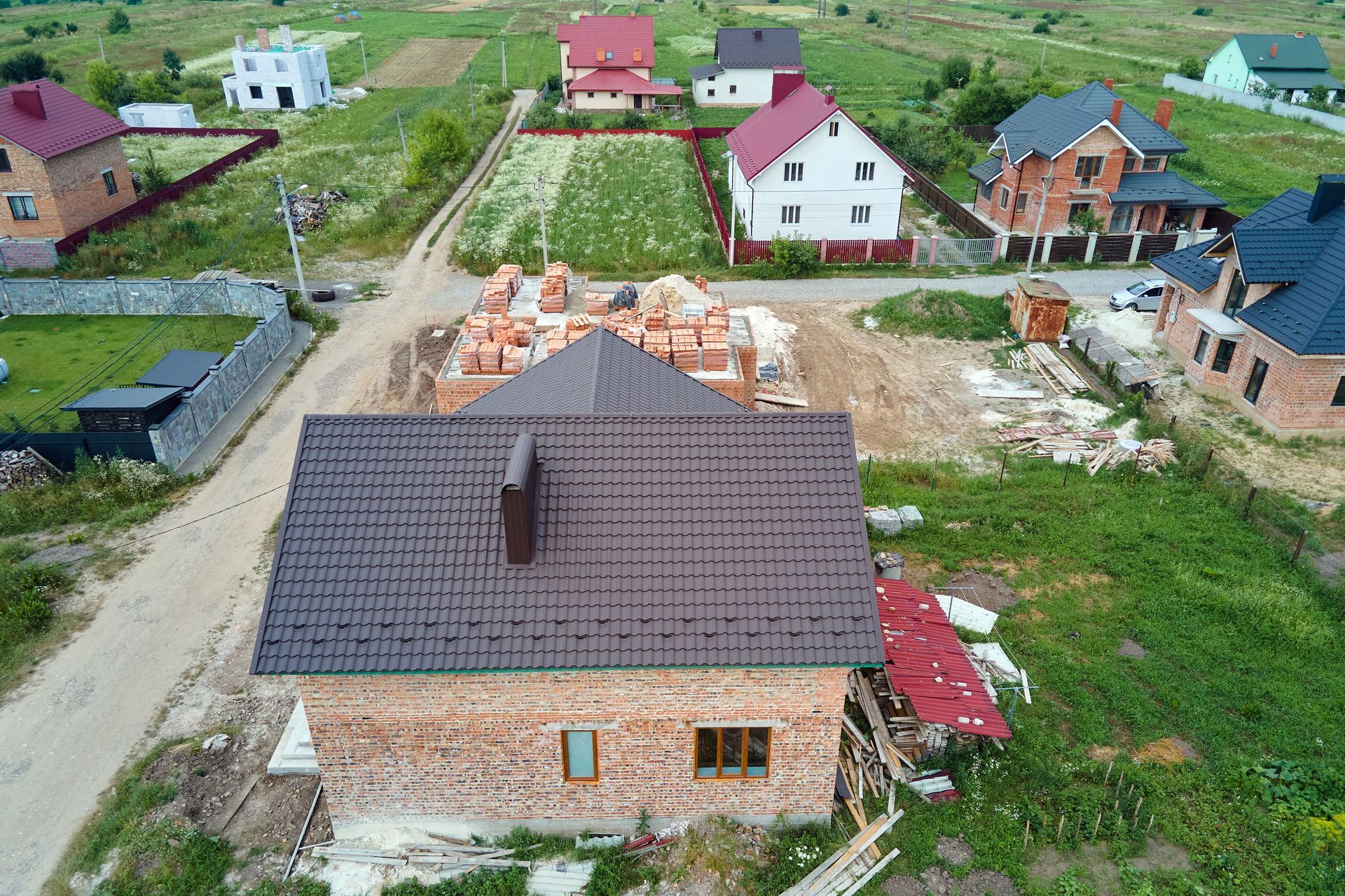 Aerial view of a brick house with a brown roof under construction, surrounded by other houses and green fields.
