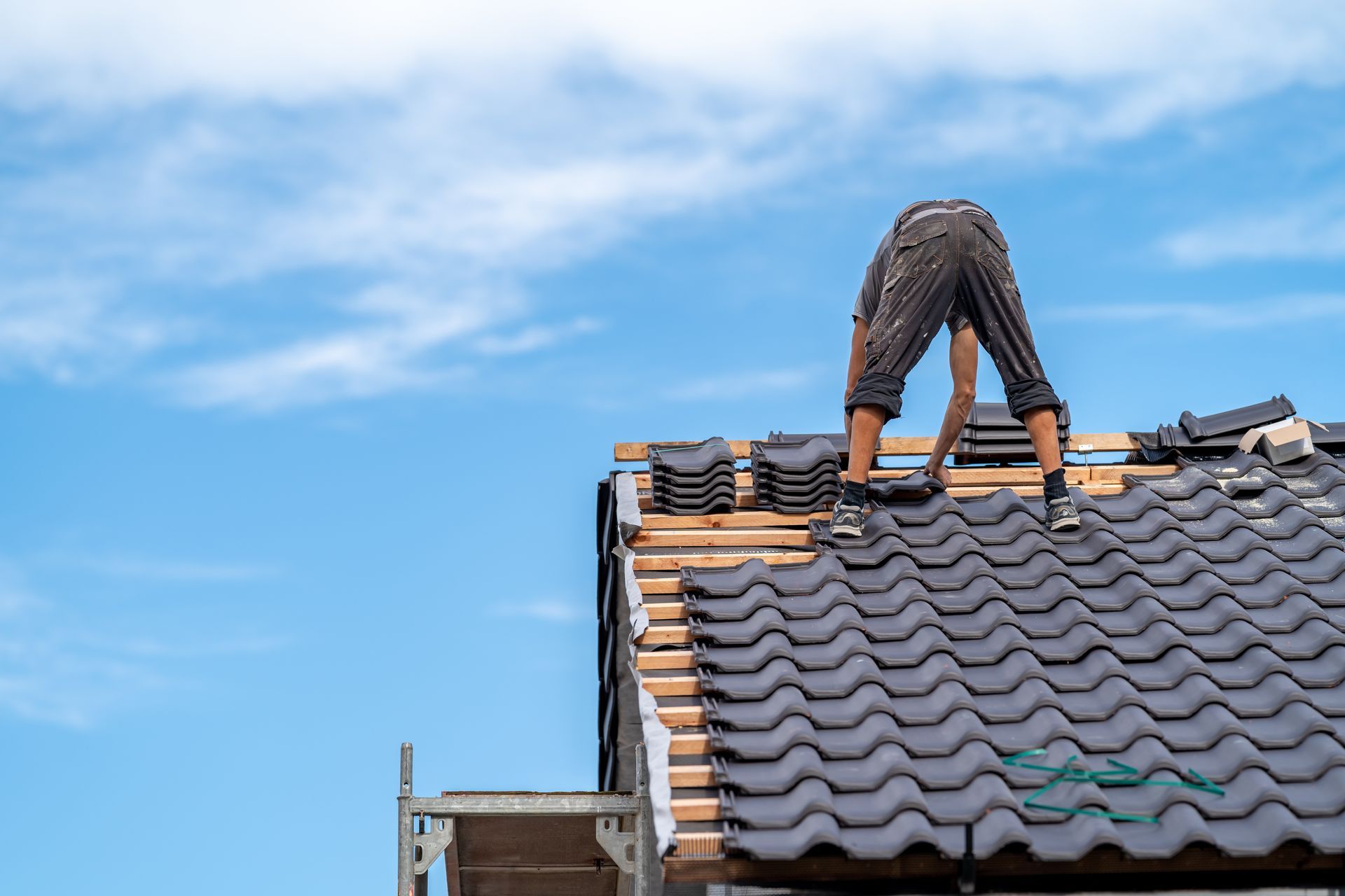 Roofer installing dark gray tiles on a roof against a bright blue sky.