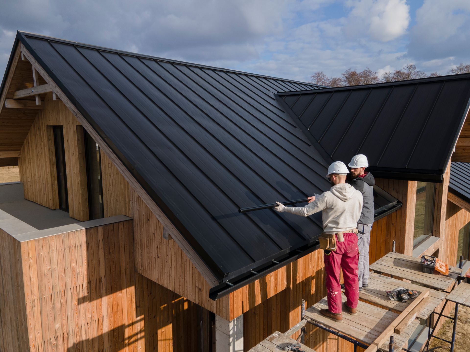 Two workers in hard hats install black metal roofing on a wooden house.