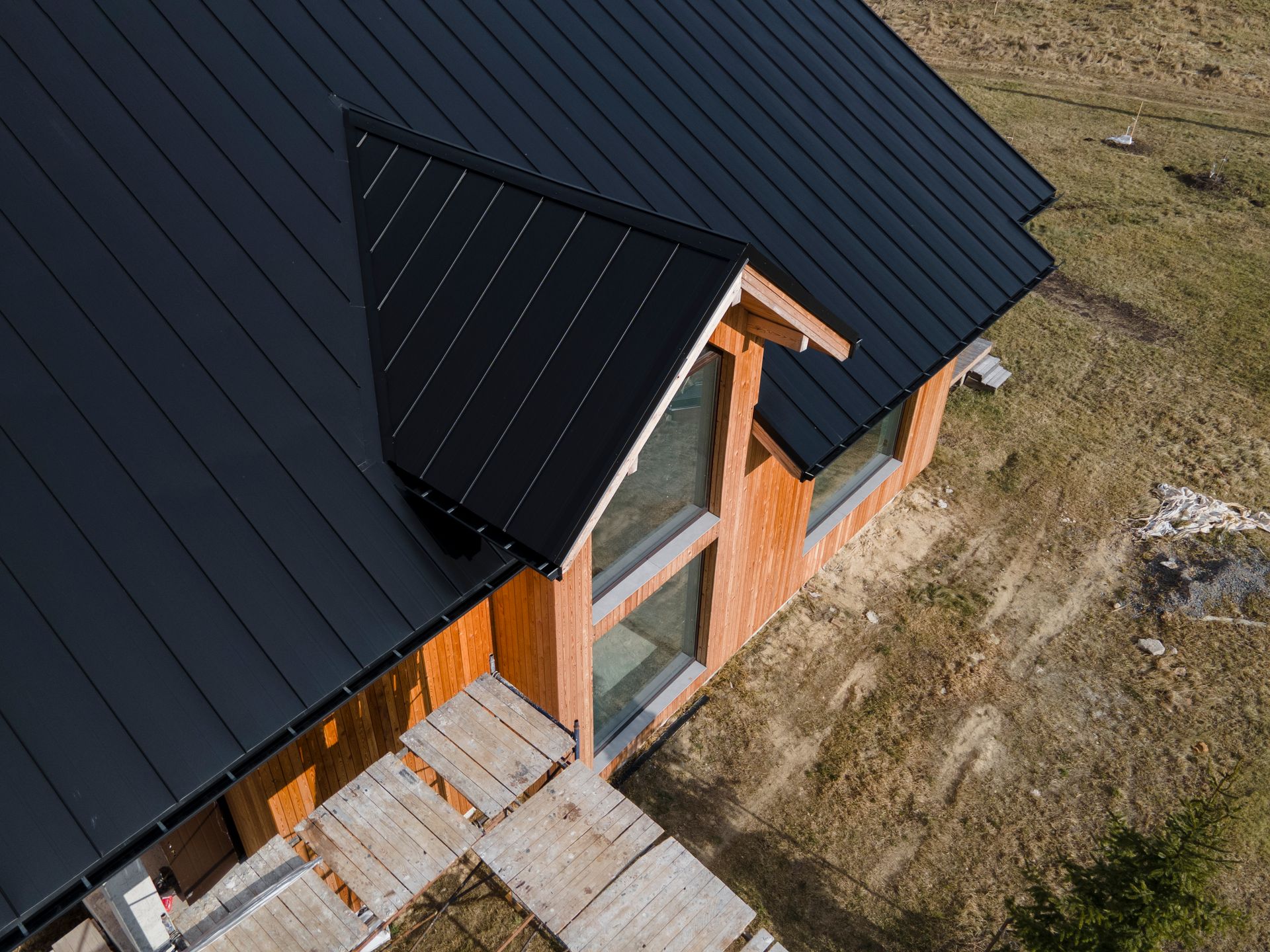 Wooden cabin with black metal roof and large windows, set in a grassy, dry landscape.
