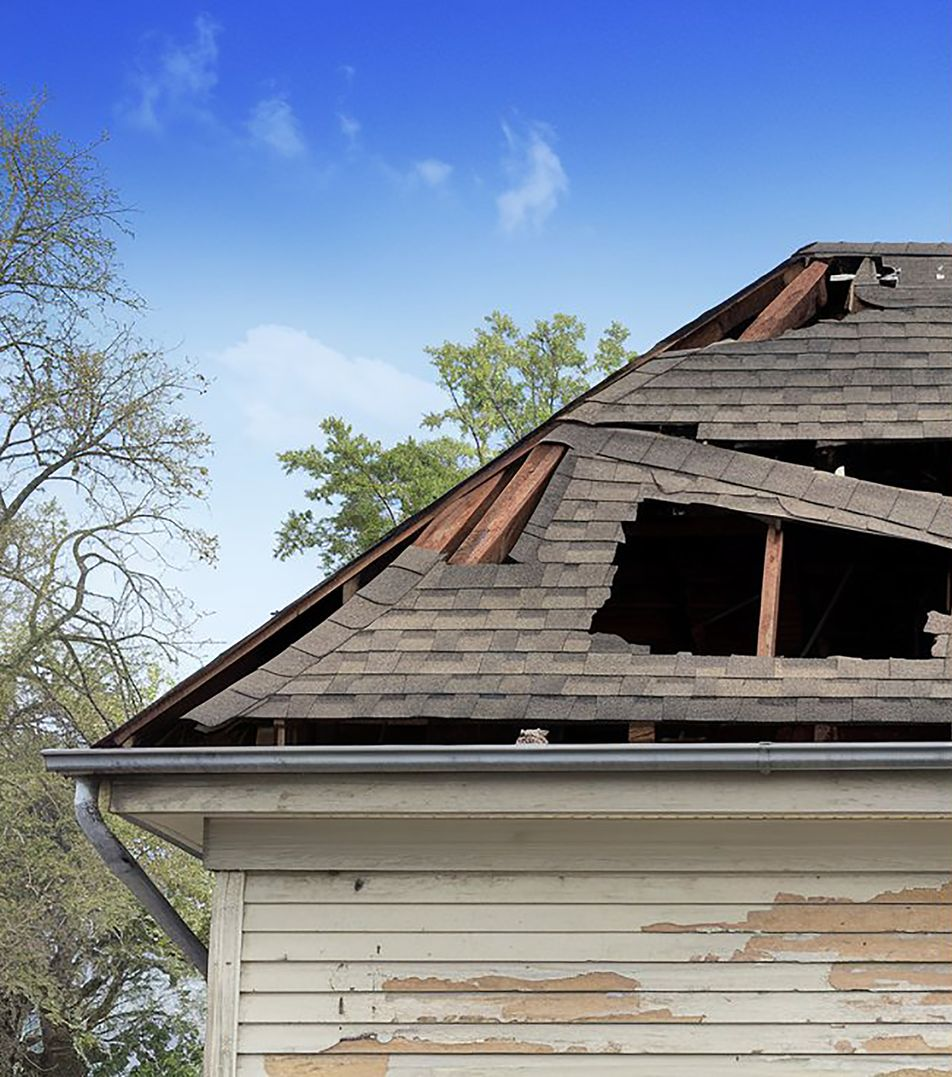 Damaged house roof with large holes, exposed wooden supports, and peeling siding against a blue sky.