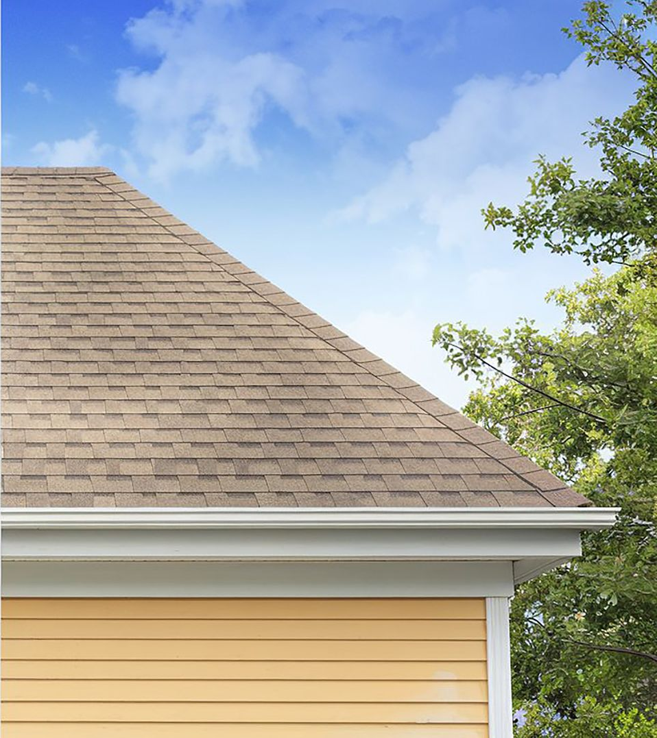 Yellow house corner with brown roof, white gutters, and green tree against a blue sky.