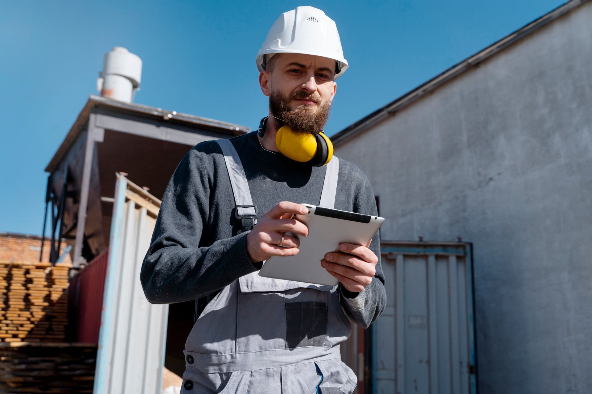 Construction worker in white hard hat and overalls using tablet outdoors.