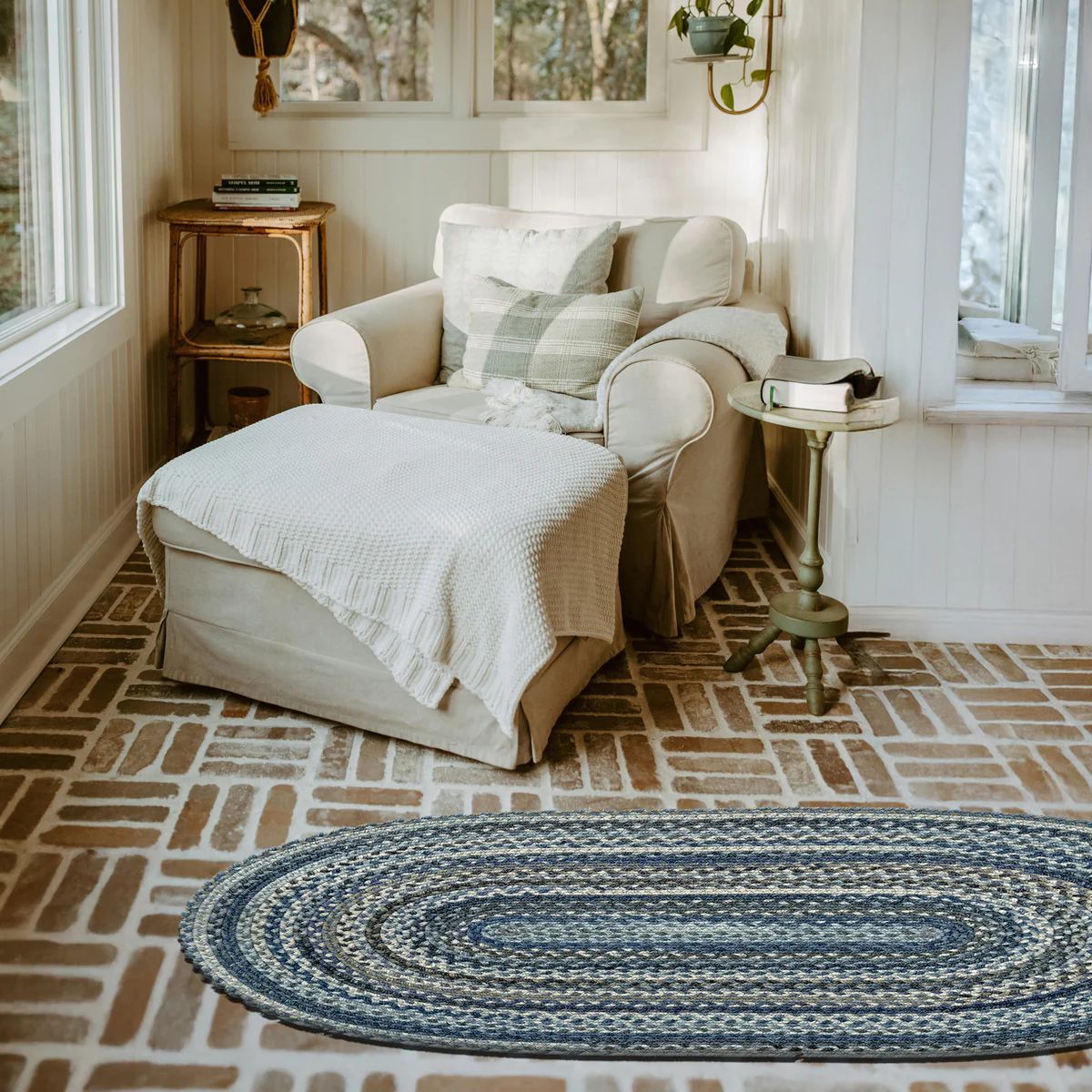 Cozy sunroom with armchair, ottoman, blue braided rug, and brick floor.