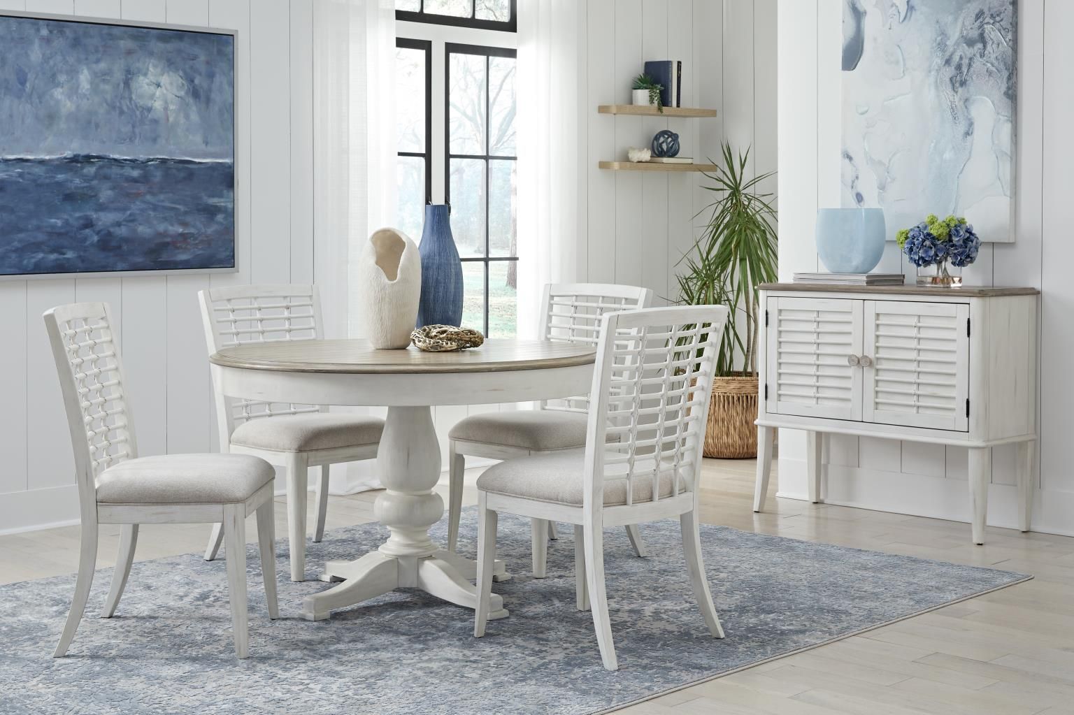 Coastal dining room: round white table with four chairs, blue rug, sideboard, art, and decor.