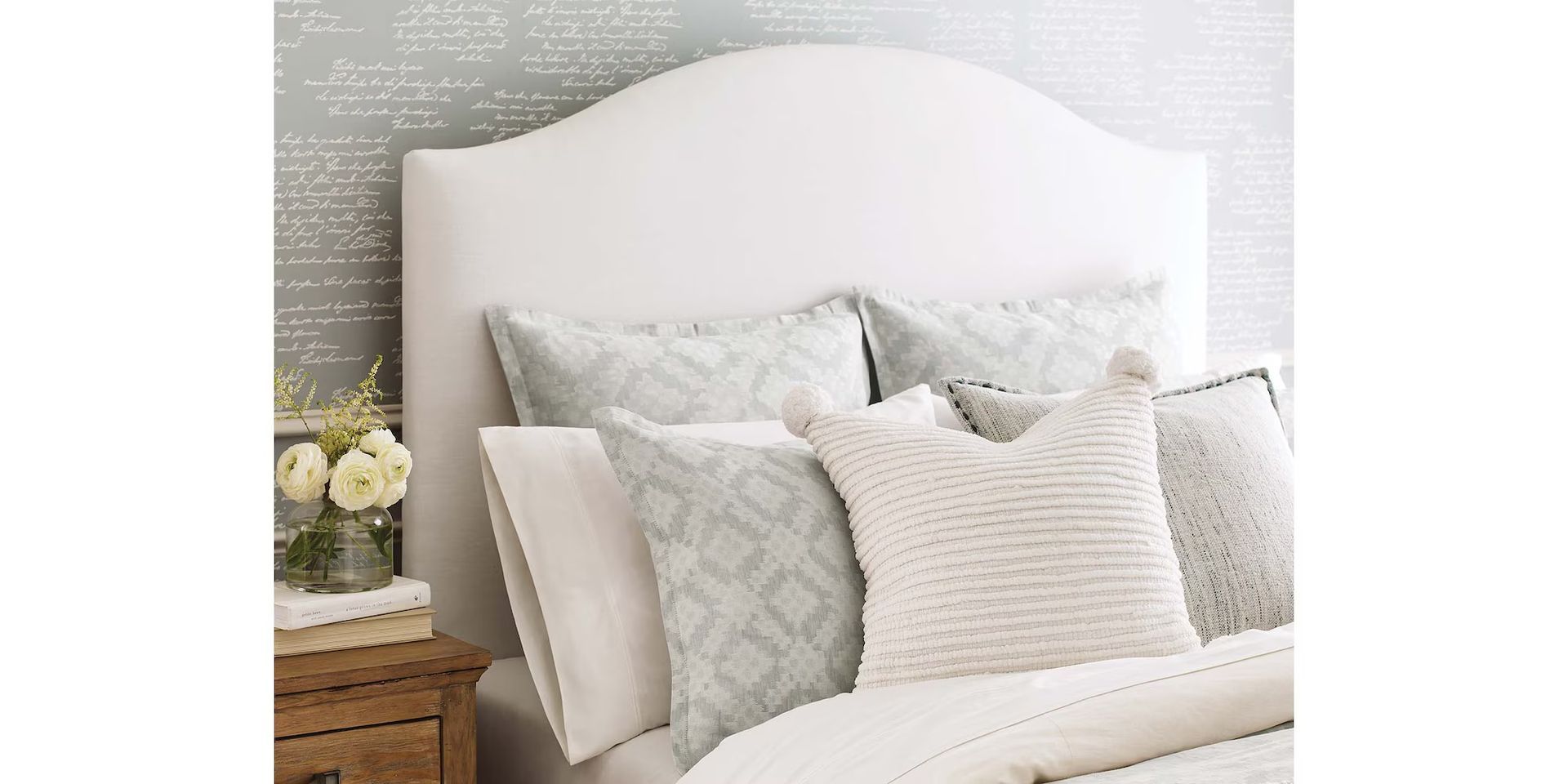 White upholstered headboard with pillows on a bed, small nightstand with flowers on the left.