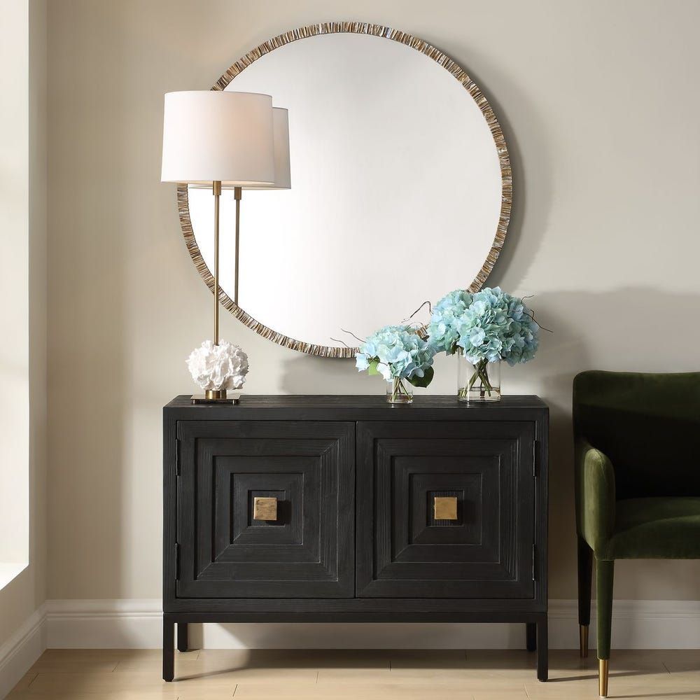 Black cabinet with gold accents, topped with a lamp and flowers, large round mirror behind.