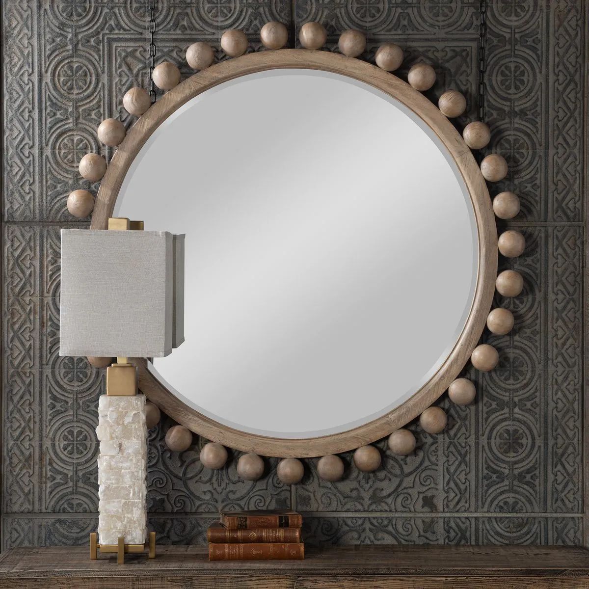Round mirror with wooden bead trim, and a lamp and books on a table.