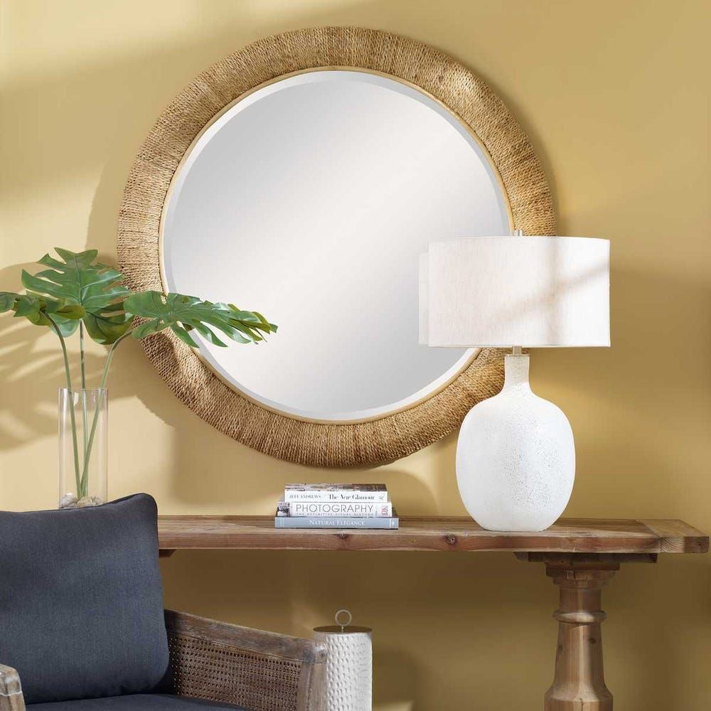 Round mirror with textured gold frame above a wooden console table. White lamp and vase with greenery on table.