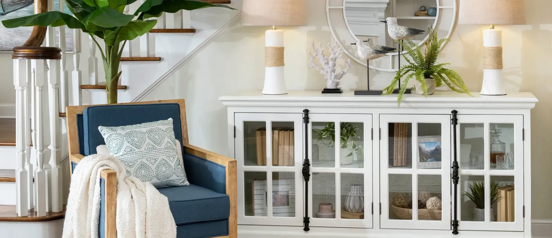 Living room with white cabinet, blue chair, lamps, and mirror. White walls and wooden staircase.