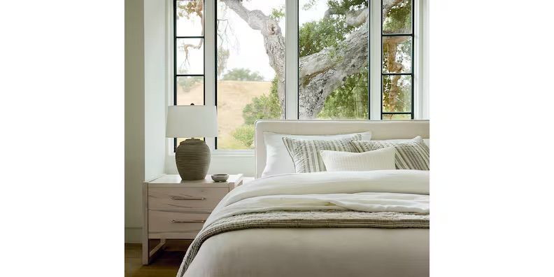 Bedroom with a neutral color palette. Bed with decorative pillows, nightstand with lamp, and large window.