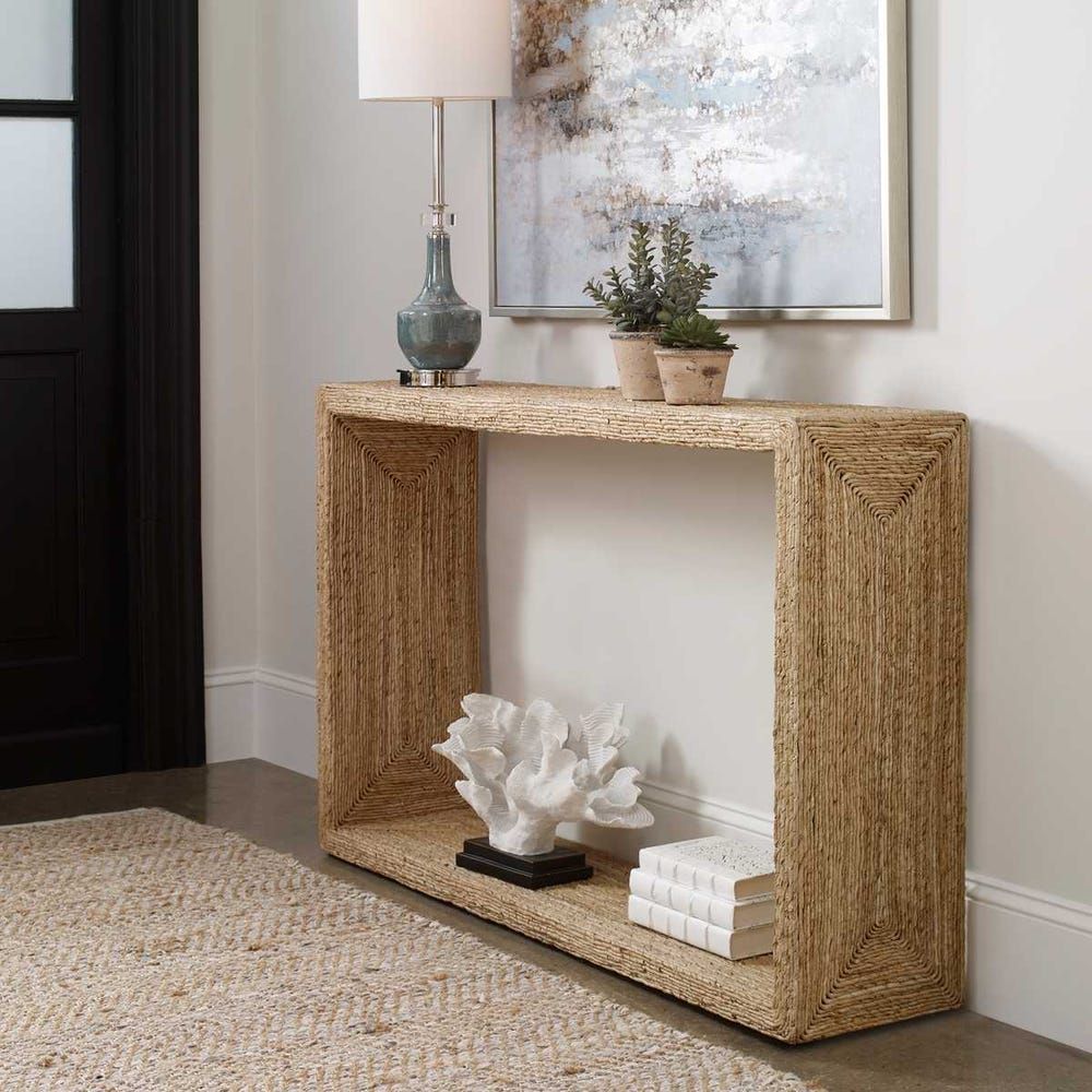 Entryway with woven jute console table, lamp, artwork, books, coral, and rug.