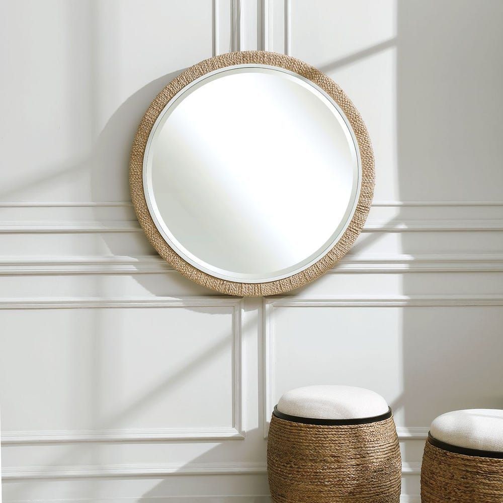 Round mirror with woven border on a white paneled wall, over two woven stools.