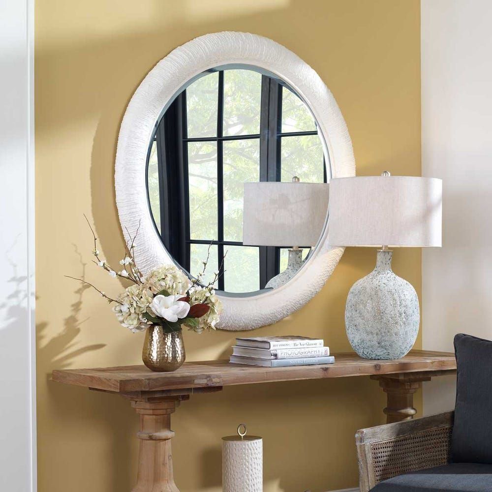 Round mirror on a yellow wall above a wood console table with a lamp, flowers, and books.