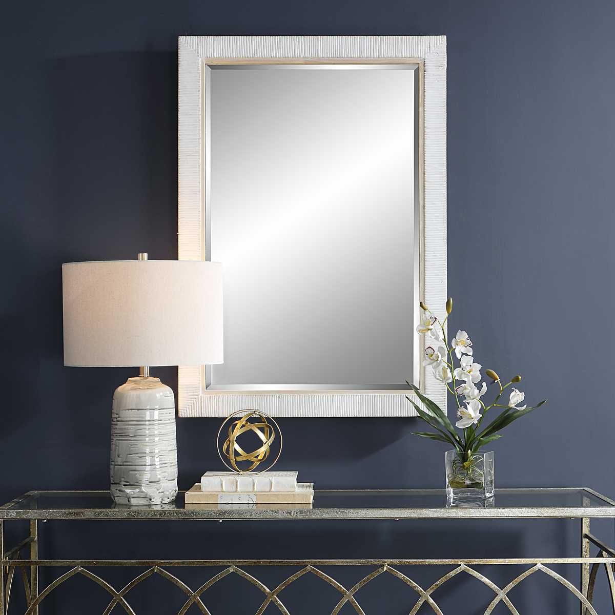 Mirror with white patterned frame, lamp, gold table, decorative ball, books, flowers on dark blue wall.