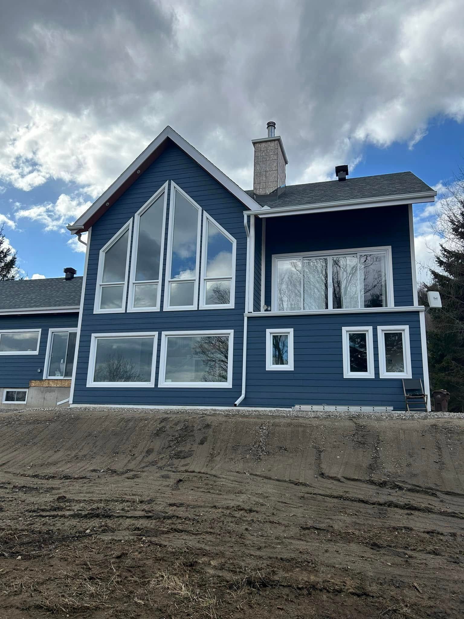 Une maison bleue à deux étages, avec de grandes fenêtres et une cheminée en pierre, sous un ciel nuageux, située sur un terrain vague non aménagé.