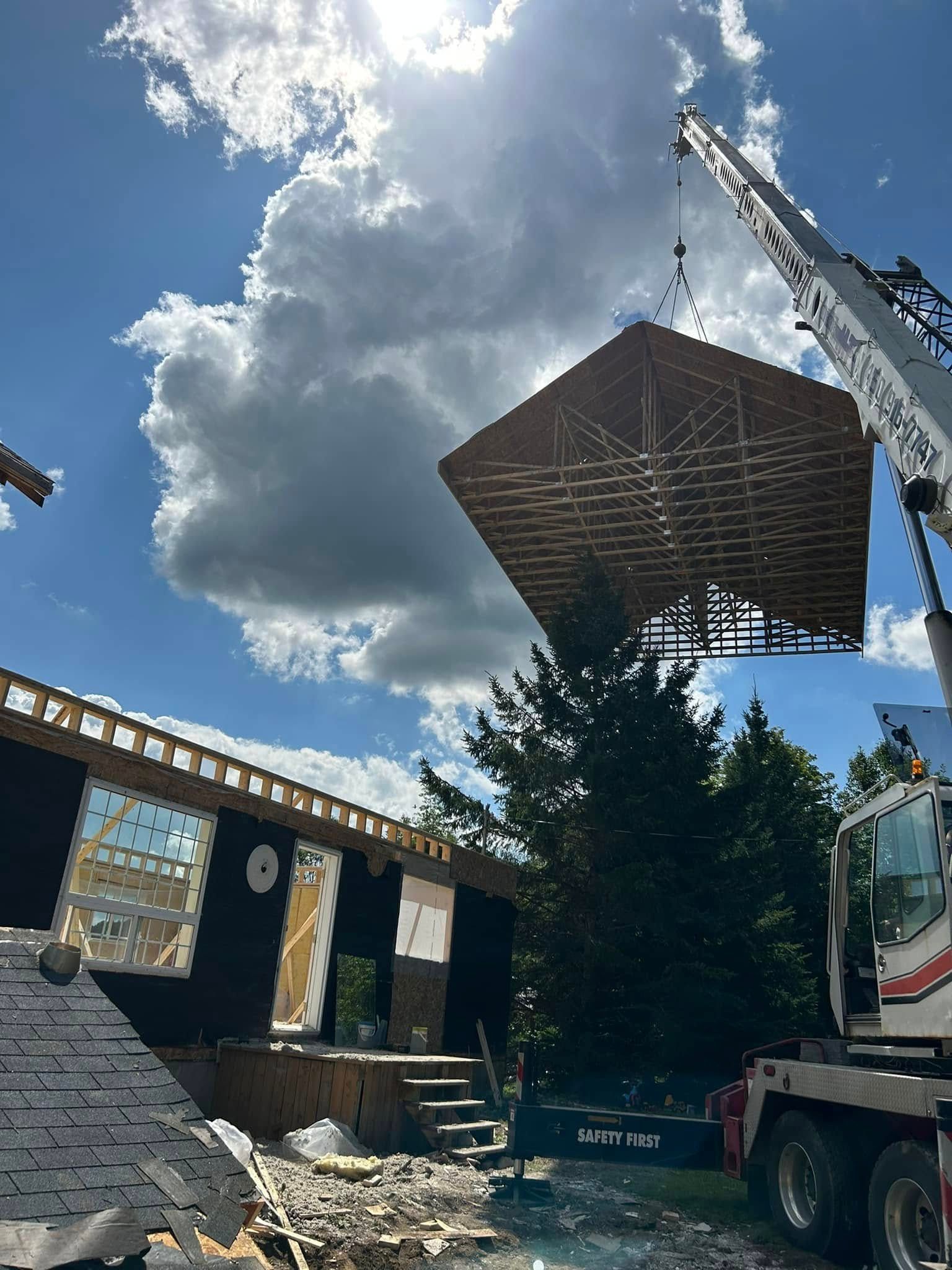 Sous un ciel bleu clair et nuageux, une grande grue soulève une charpente de toit en bois et la met en place au-dessus de la structure du bâtiment.