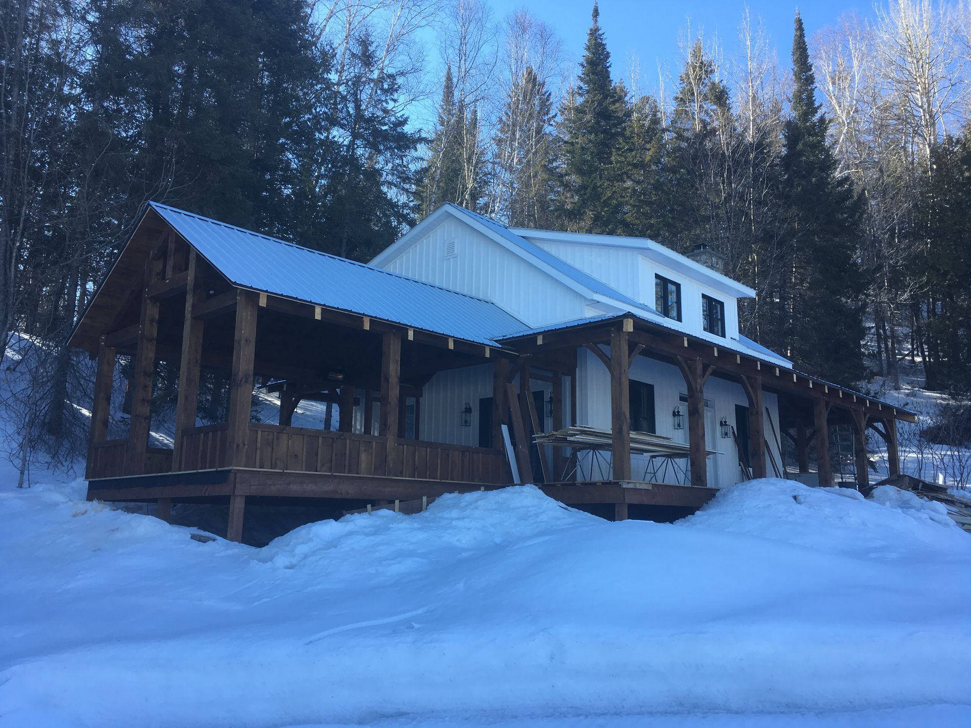 Une cabane avec une vaste véranda en bois recouverte de neige, entourée d'une forêt dense par une journée d'hiver ensoleillée